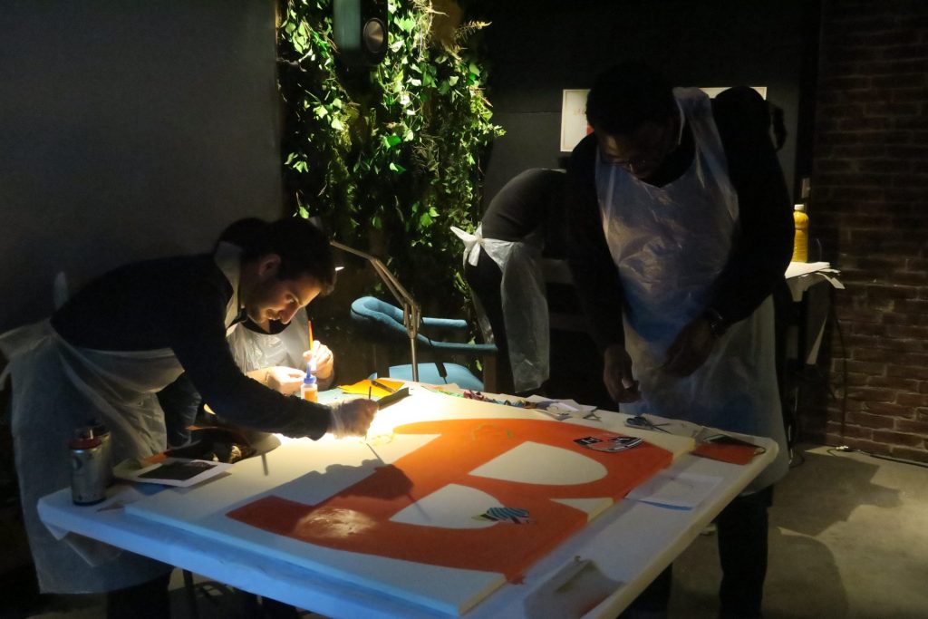 Deux participants en tablier peignent une grande toile orange pendant l’événement Hermès au Kube Hôtel, organisé par Weevup