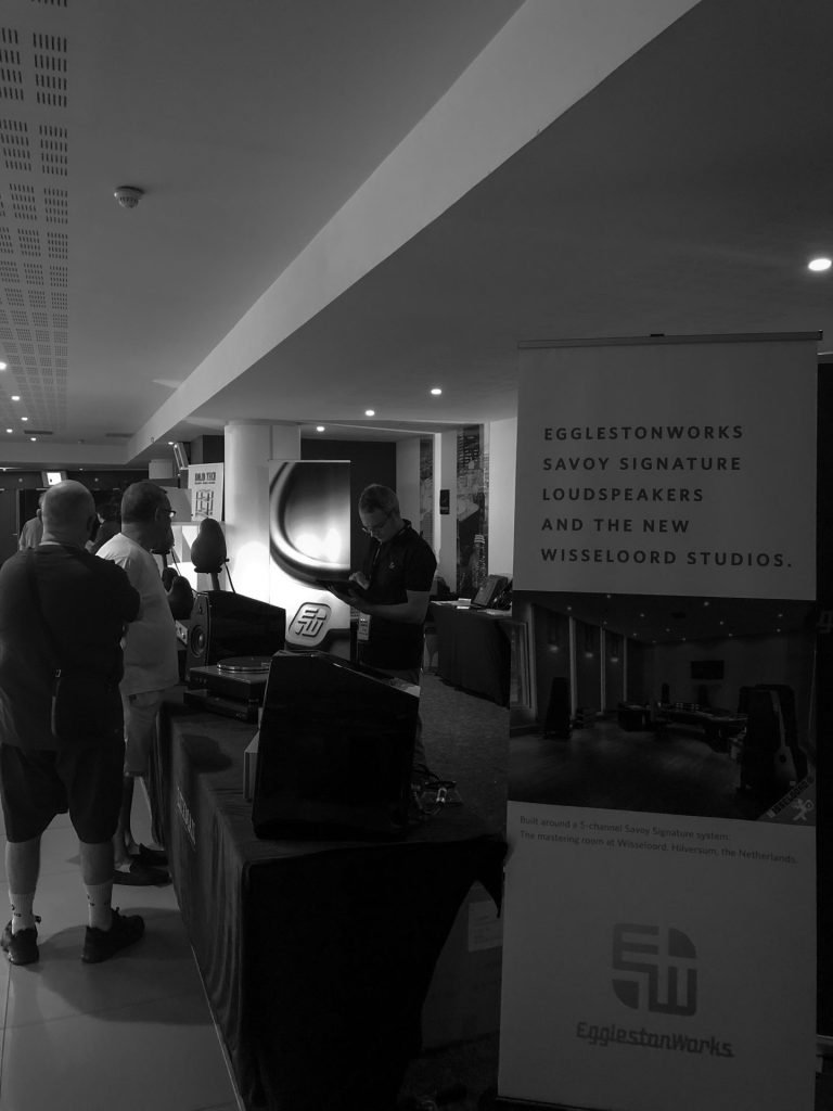 Visiteurs devant un stand d’enceintes au salon Publison, au Novotel Paris Est, avec un exposant derrière la table