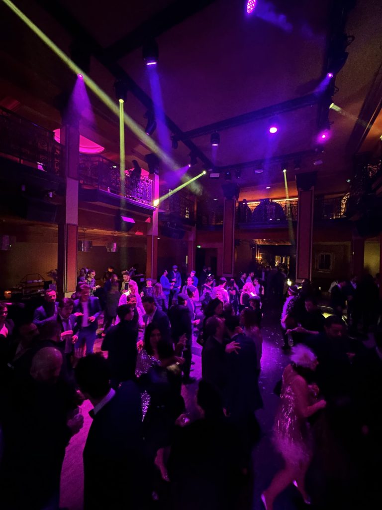 Invitados de Scalian bailando bajo haces de luz verde y violeta, algunos con vestidos de los años veinte, tras los premios de su velada temática Gatsby en un antiguo banco