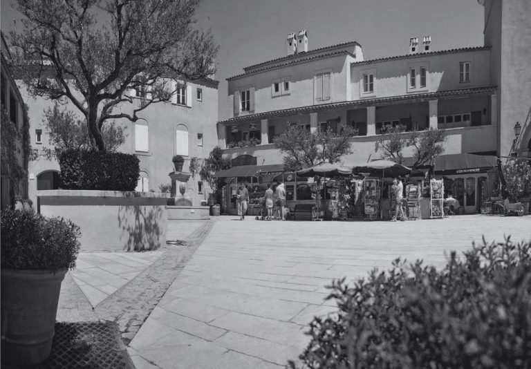 Place d’un village provençal, avec ses façades claires, ses oliviers et ses boutiques, en noir et blanc