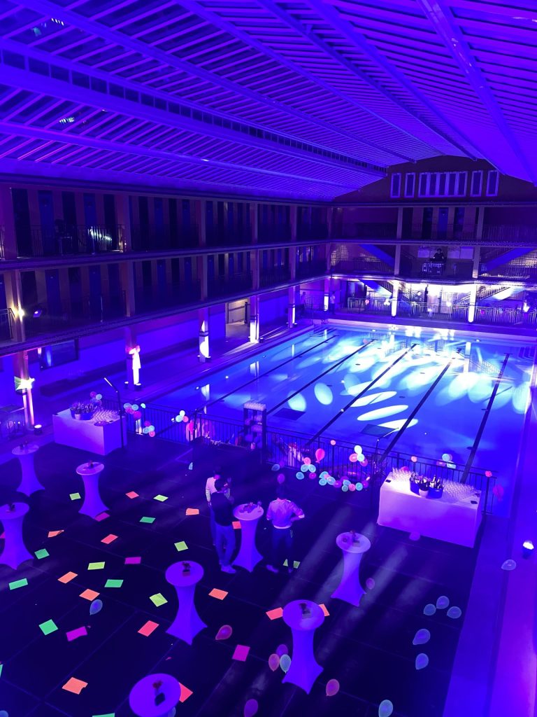 Scénographie fluo vue d’en haut, avec carrés colorés au sol, mange-debout, ballons et bars au bord du bassin, soirée Havea Group organisée par Weevup