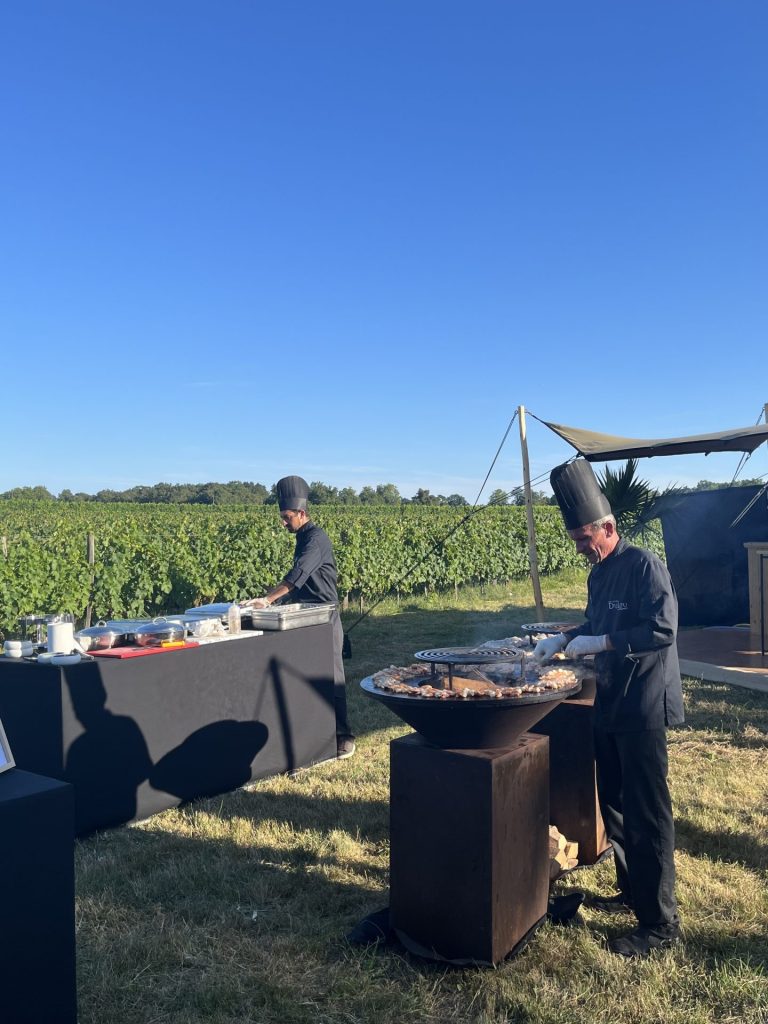 Deux cuisiniers en toque, l’un au brasero, l’autre à sa table de préparation, face aux vignes, pendant un séminaire au château Lafitte