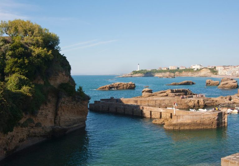 Falaise verdoyante, rochers et jetées en pierre dans une mer turquoise, avec un phare blanc au loin
