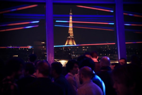 Une foule de personnes dans un club ou un bar, avec en arrière-plan la Tour Eiffel illuminée. Des lumières horizontales traversent la scène, créant une ambiance festive. La scène capture une soirée animée avec la silhouette emblématique de la Tour Eiffel dominant l'arrière-plan. convention outscale 3ds