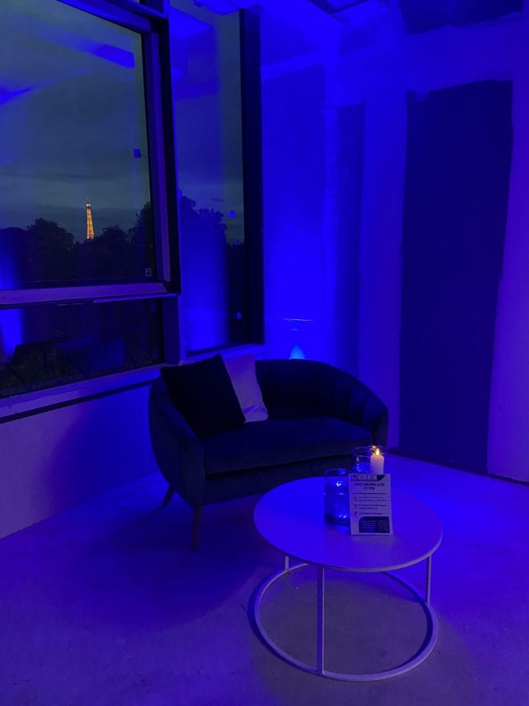 Coin salon avec fauteuil et table basse sous des lumières bleues, devant une fenêtre où l’on aperçoit la tour Eiffel au loin, pendant la soirée Delpha organisée par Weevup sur un chantier