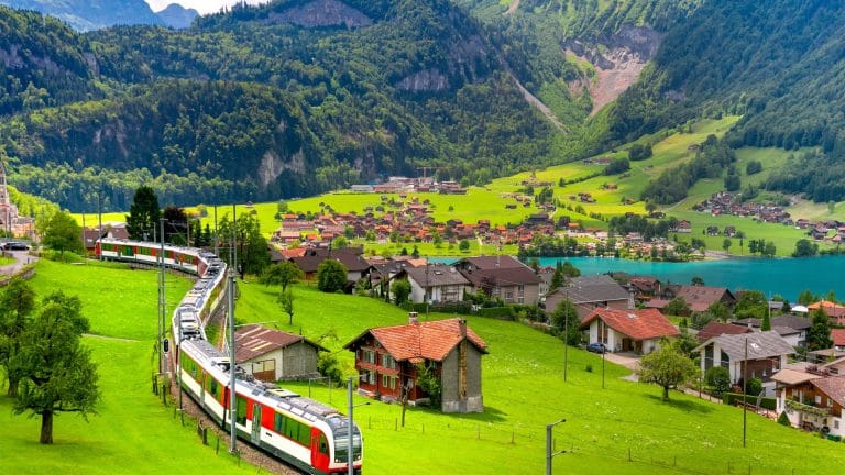 Train rouge et blanc traversant une vallée alpine en Suisse, entre chalets, prairies et lac turquoise