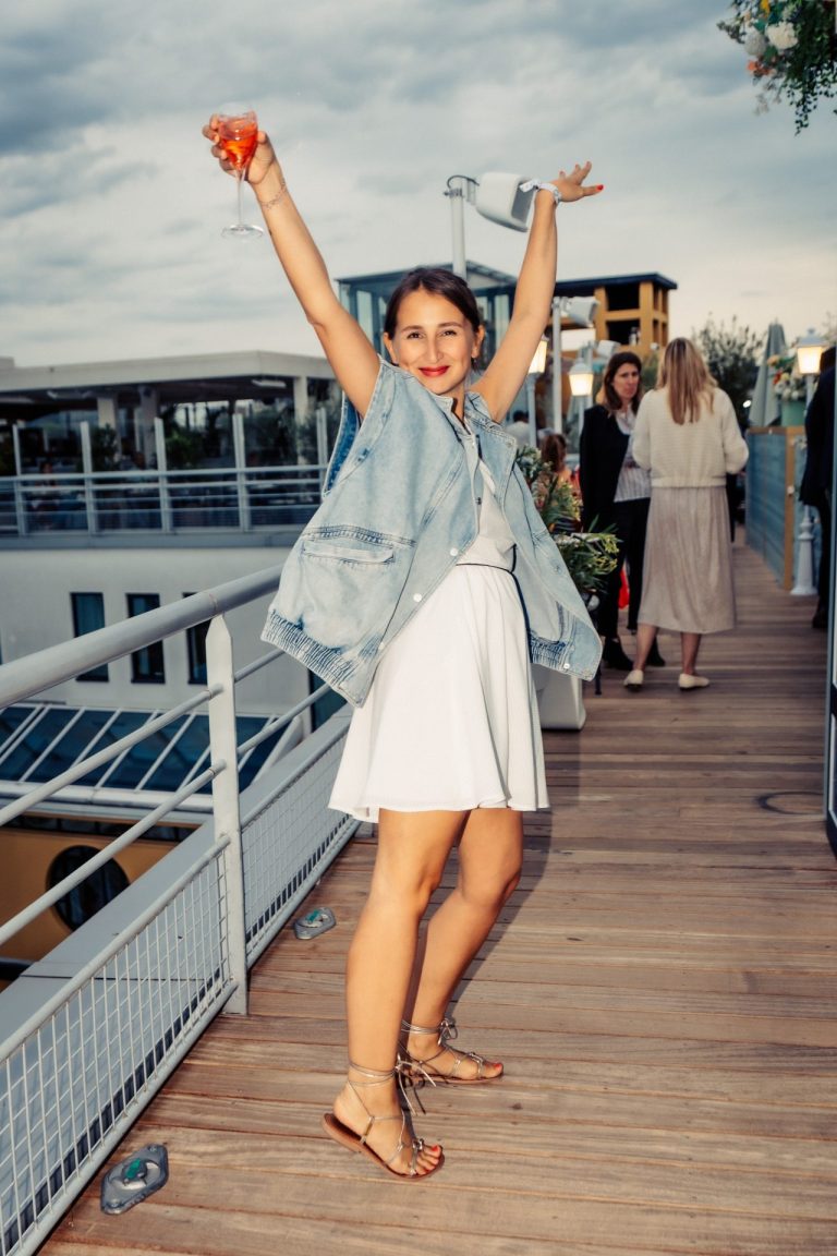 Invitée levant les bras, un verre à la main, sur le rooftop au plancher de bois de la cocktail party d’été de Dior organisée par Weevup