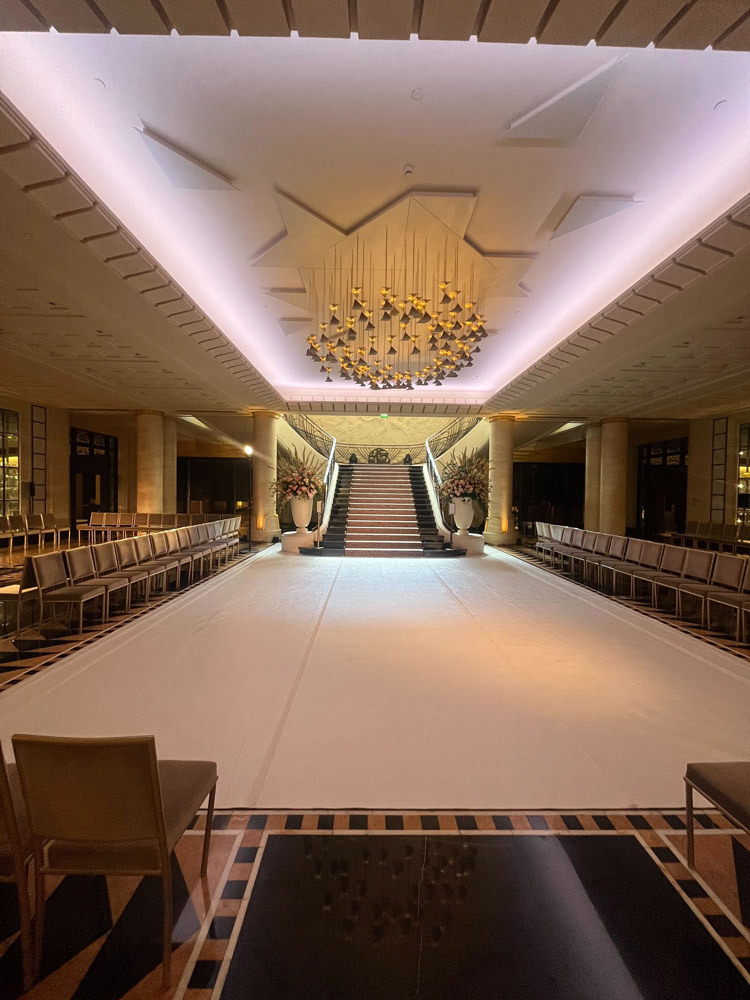 Podium blanc bordé de chaises devant un escalier, sous un lustre, prêt pour le défilé Yara Shoemaker à l’Hôtel Le Collectionneur à Paris