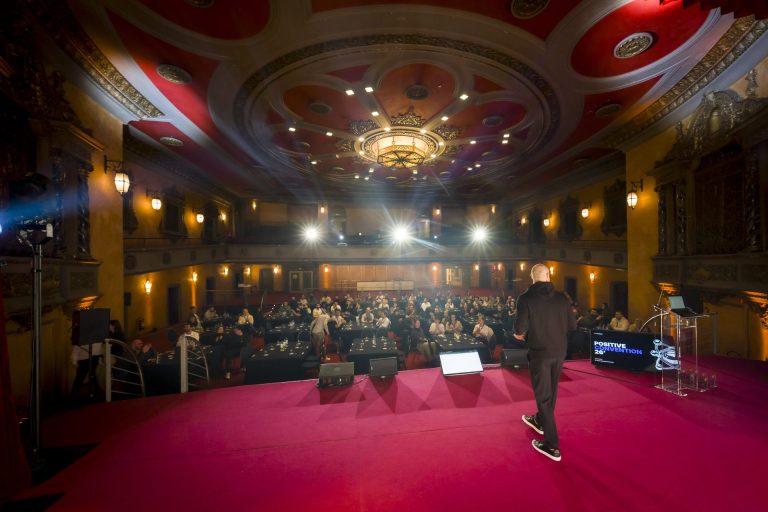 Orateur sur scène face au public de la convention Groupe Positive, dans une salle de théâtre à Bruxelles
