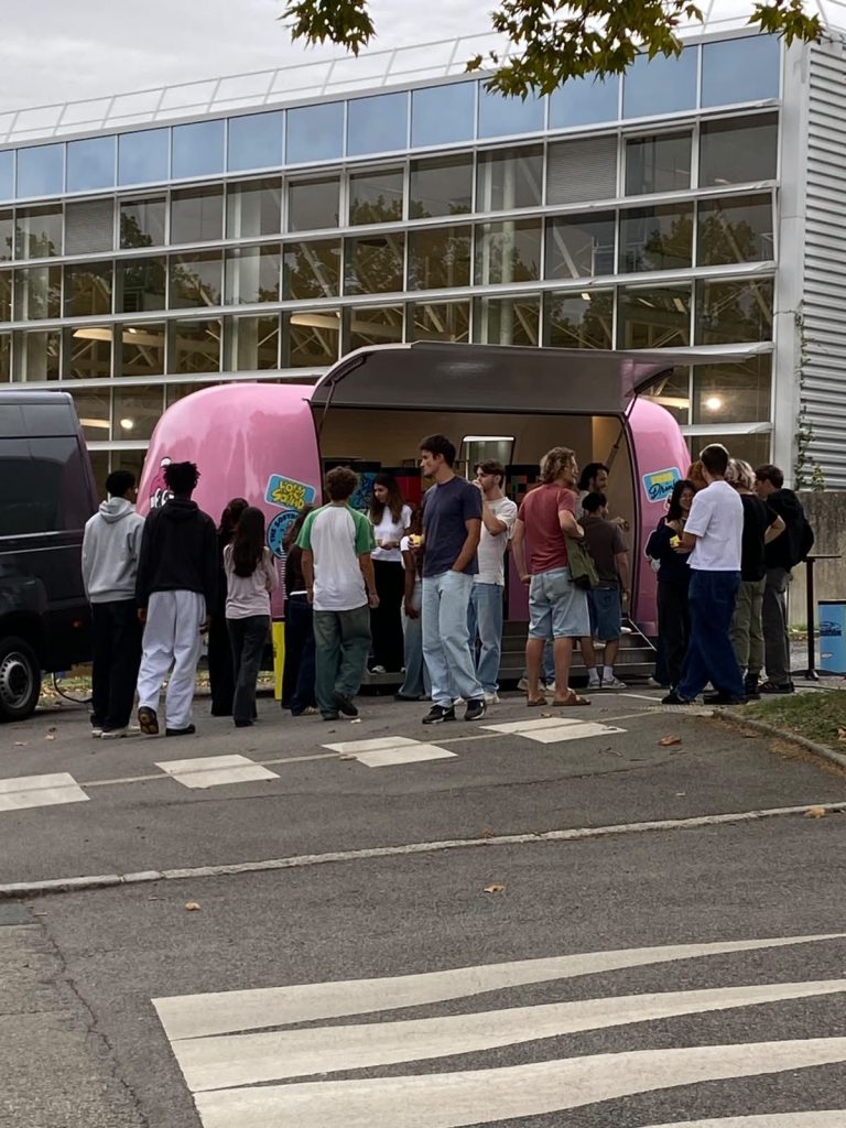 Remorque-bar rose du Holy Campus Tour ouverte devant un bâtiment de campus, une foule réunie autour pour la dégustation