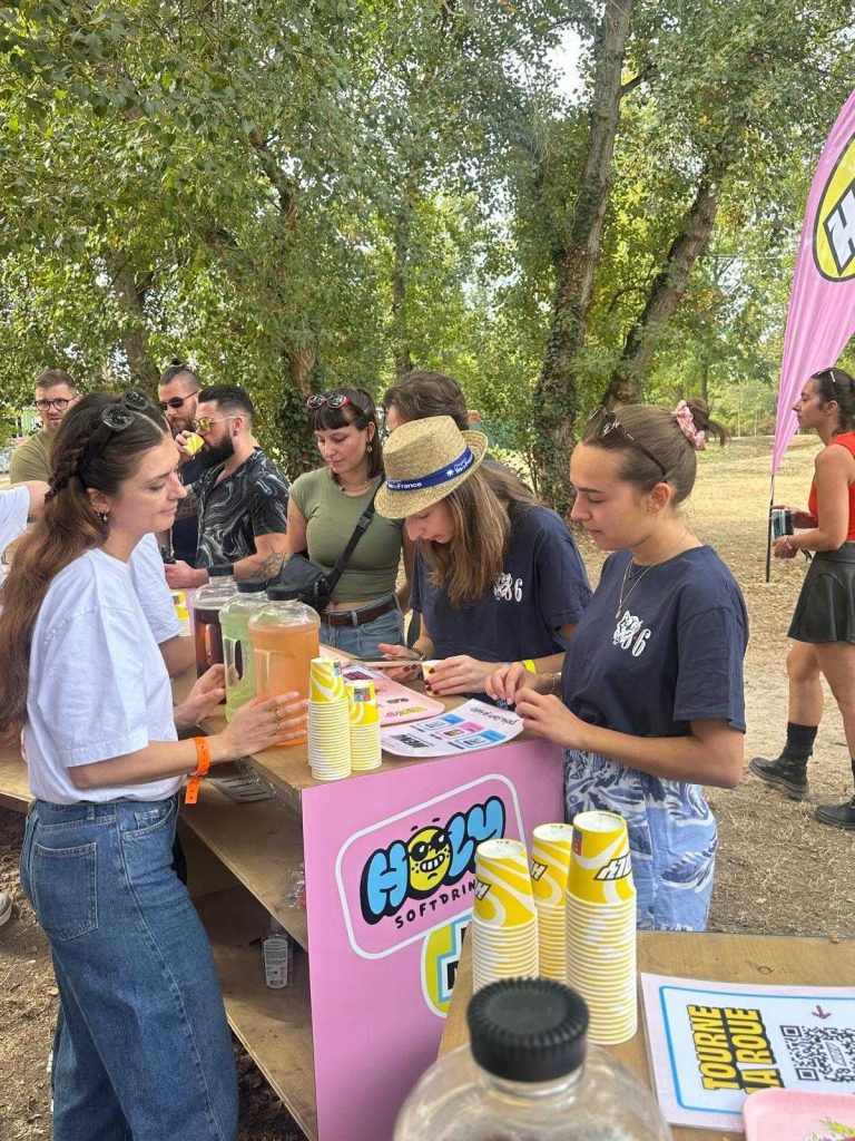 Festivaliers au comptoir de dégustation Holy à Elektric Park, gobelets jaunes et carafes de boissons sur le bar