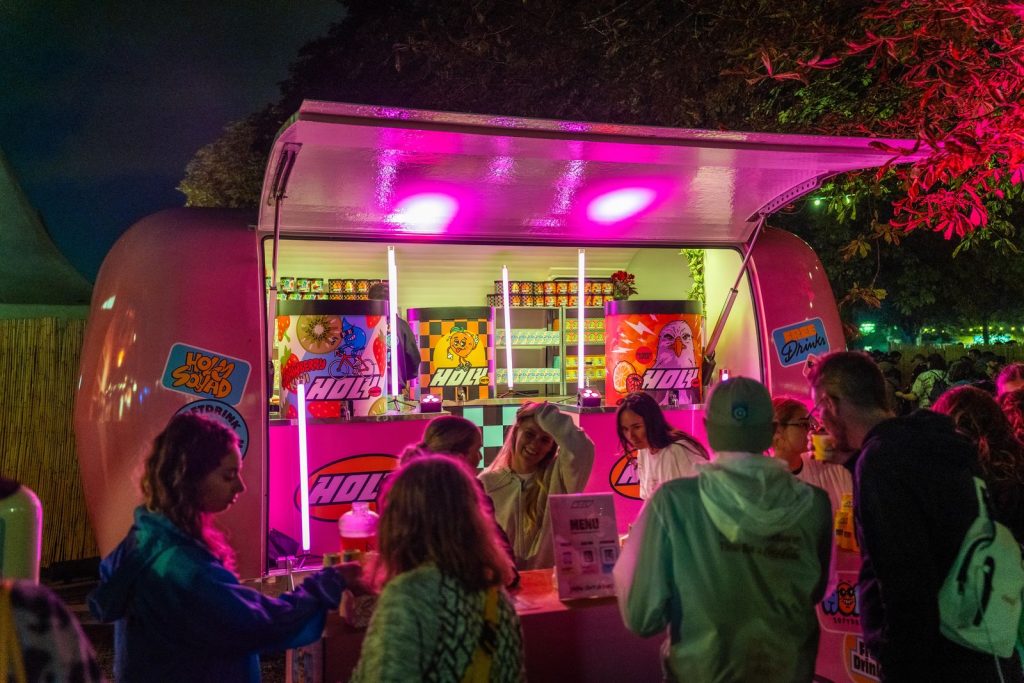 La caravane Holy éclairée de néons roses à la nuit tombée, festivaliers au comptoir de dégustation