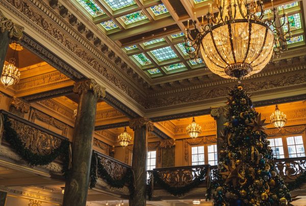Grand sapin de Noël décoré de boules dorées et bleues dans un hall de prestige, sous une verrière colorée et des lustres en cristal, avec des guirlandes de verdure aux balcons