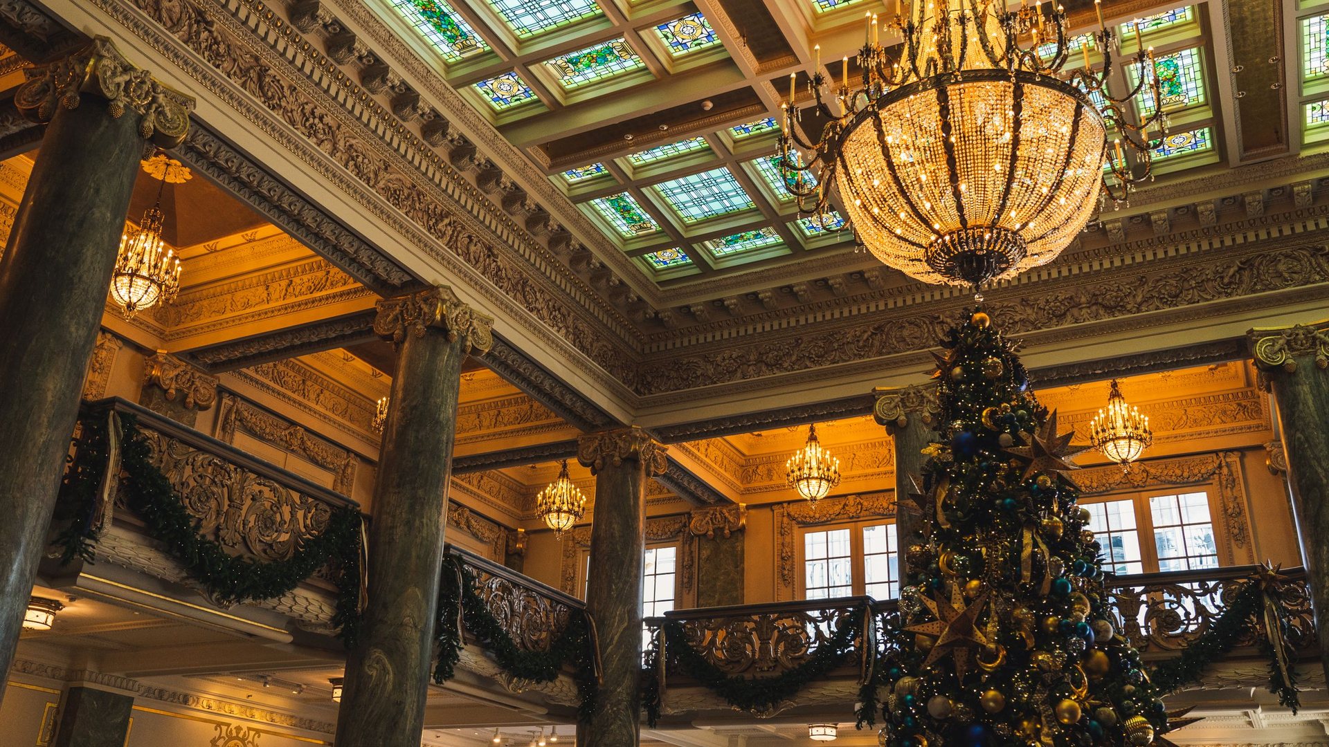 Grand sapin de Noël décoré de boules dorées et bleues dans un hall de prestige, sous une verrière colorée et des lustres en cristal, avec des guirlandes de verdure aux balcons