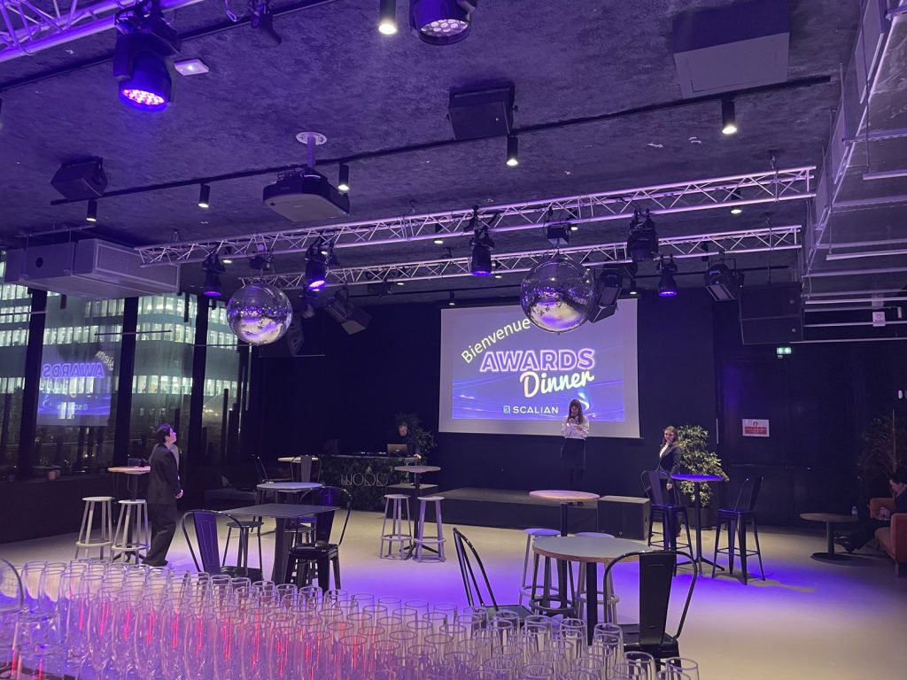 Salle du Nodd avant l’arrivée des invités : écran « Bienvenue Awards Dinner Scalian », boules à facettes, mange-debout et rangées de flûtes, immeubles de bureaux éclairés derrière la baie vitrée