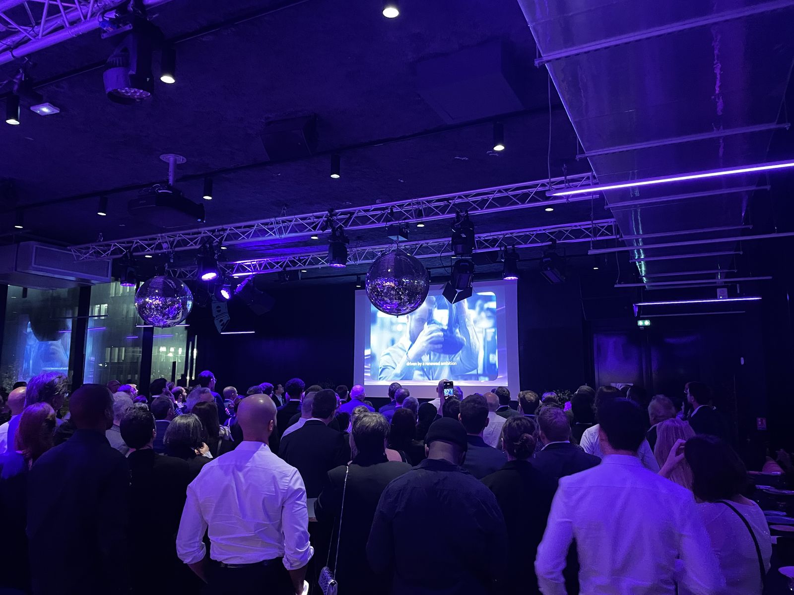 Invités de l'Awards Dinner Scalian debout face au grand écran du Nodd pendant la diffusion d'une vidéo, sous les projecteurs violets et les boules à facettes.