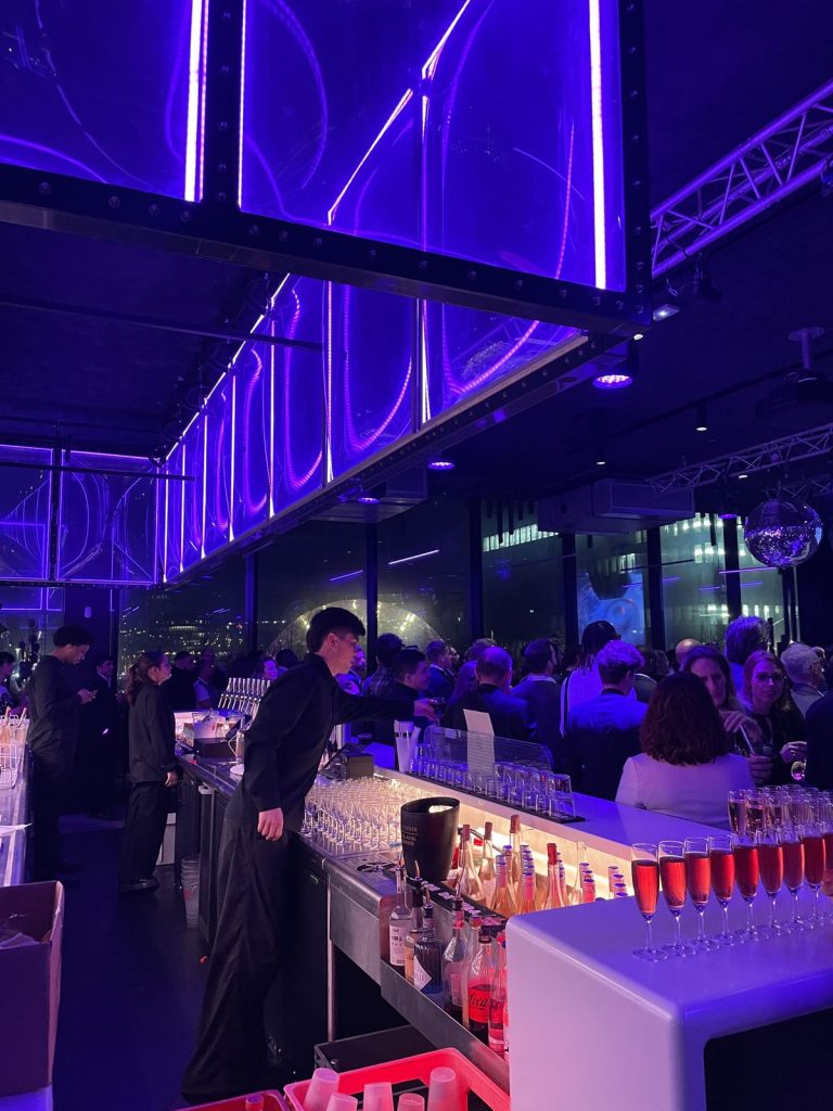 Bar du Nodd pendant l’Awards Dinner Scalian : flûtes de kir alignées sur le comptoir éclairé, barman au service et invités sous les caissons lumineux violets