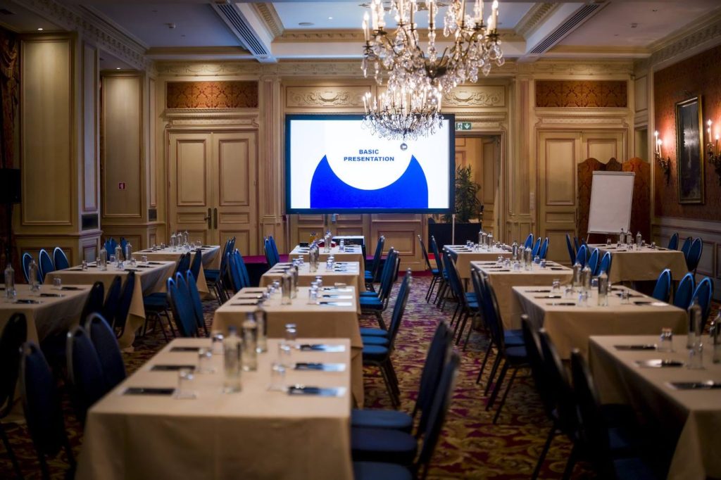Salle de réunion sous un grand lustre en cristal, tables nappées et chaises bleues face à un écran, préparée pour la convention du Groupe Positive à Bruxelles organisée par Weevup