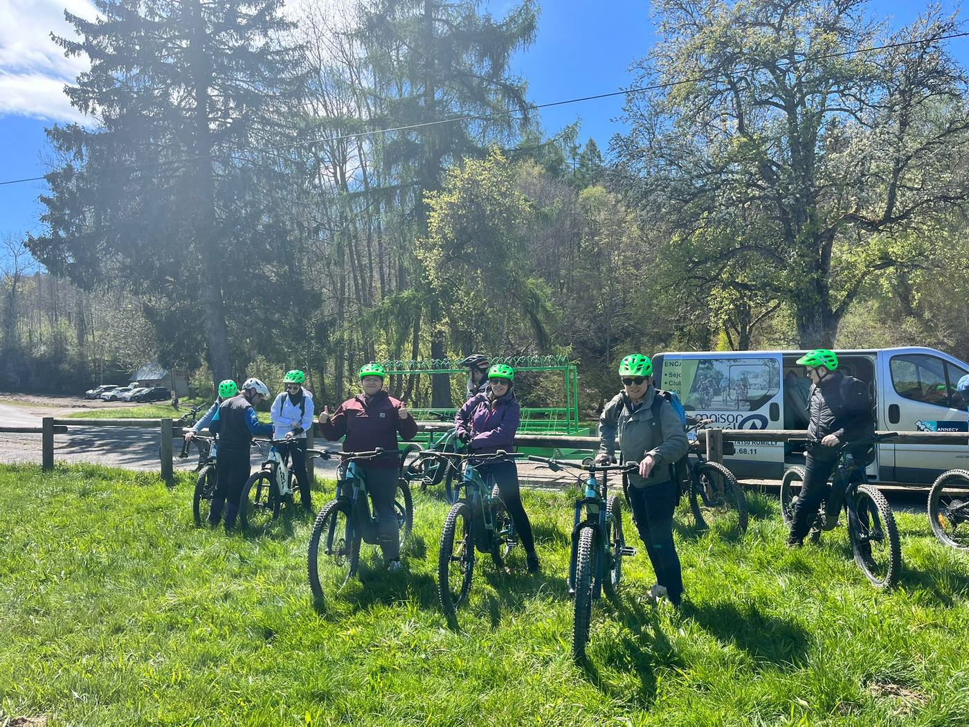 Participants en casques verts prêts à partir à vélo électrique, sur une pelouse ensoleillée en lisière de forêt