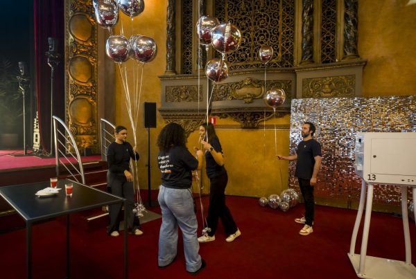 Équipe Weevup installant des ballons argentés et une borne photo au pied de la scène, dans une salle de spectacle aux ornements dorés et à la moquette rouge, pendant la préparation de la convention du Groupe Positive à Bruxelles