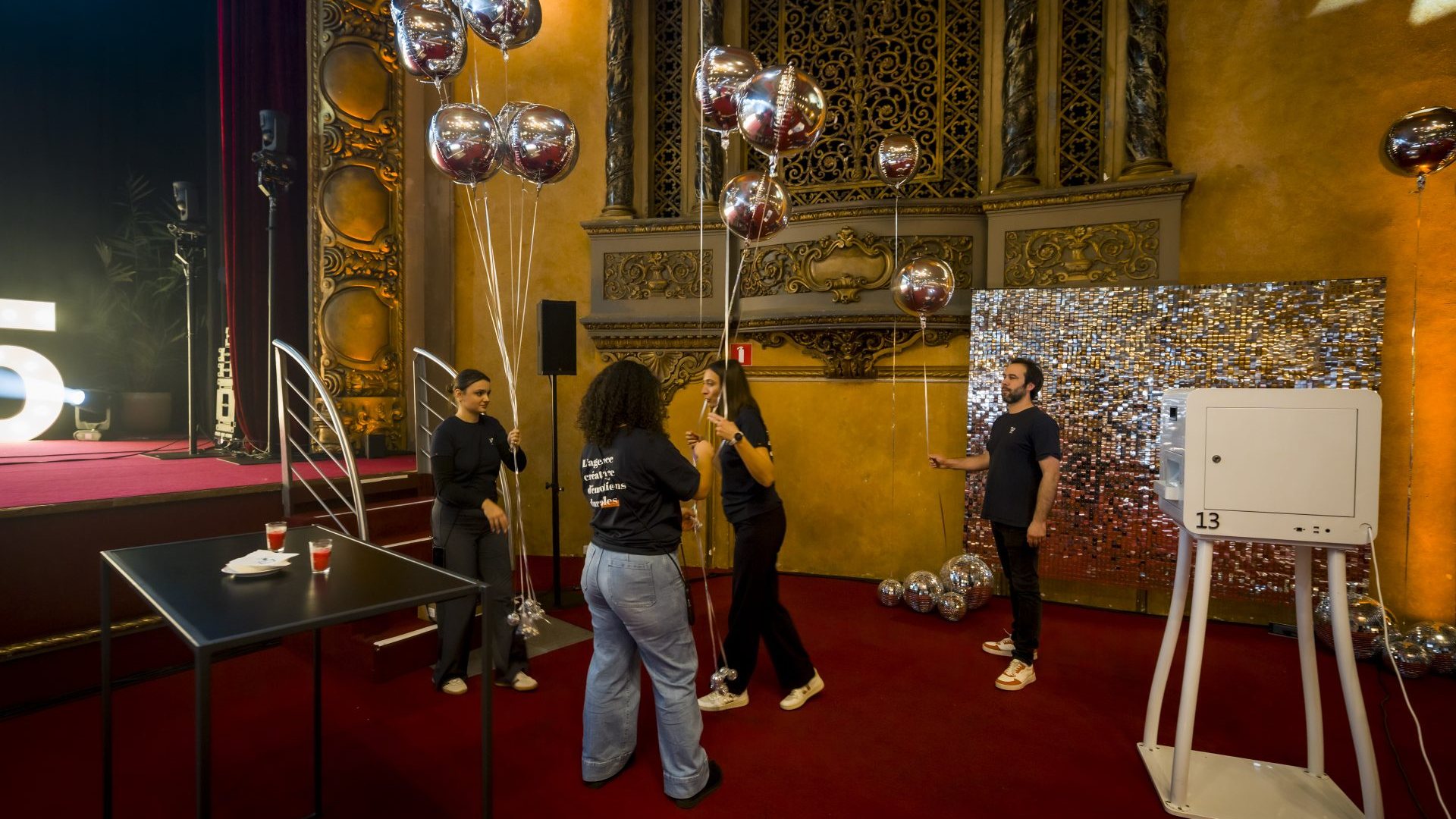 Équipe Weevup installant des ballons argentés et une borne photo au pied de la scène, dans une salle de spectacle aux ornements dorés et à la moquette rouge, pendant la préparation de la convention du Groupe Positive à Bruxelles