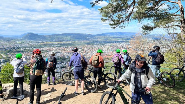 Groupe en sortie VTT sur un belvédère, face au panorama d’une ville entourée de montagnes