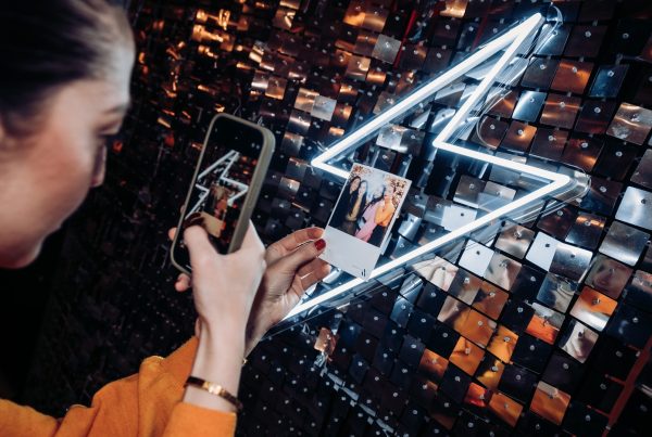 Invitée photographiant avec son téléphone son tirage photo souvenir devant un mur de sequins et un éclair en néon, lors de la soirée de lancement de Vanessa Wu organisée par Weevup