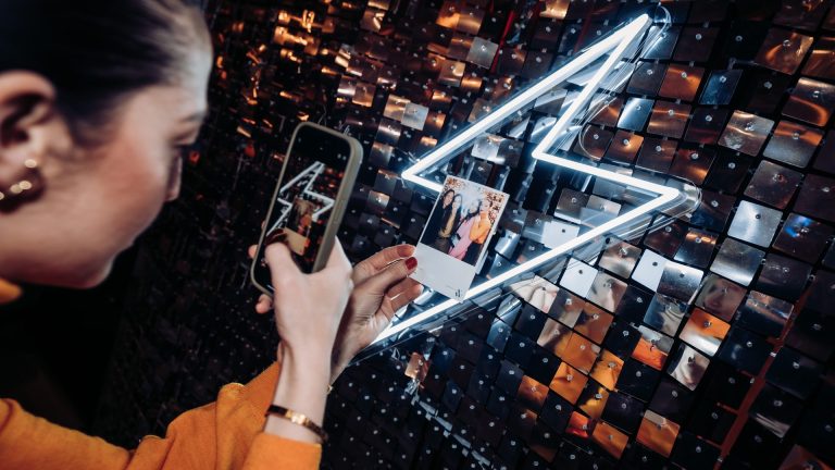 Invitée photographiant avec son téléphone son tirage photo souvenir devant un mur de sequins et un éclair en néon, lors de la soirée de lancement de Vanessa Wu organisée par Weevup
