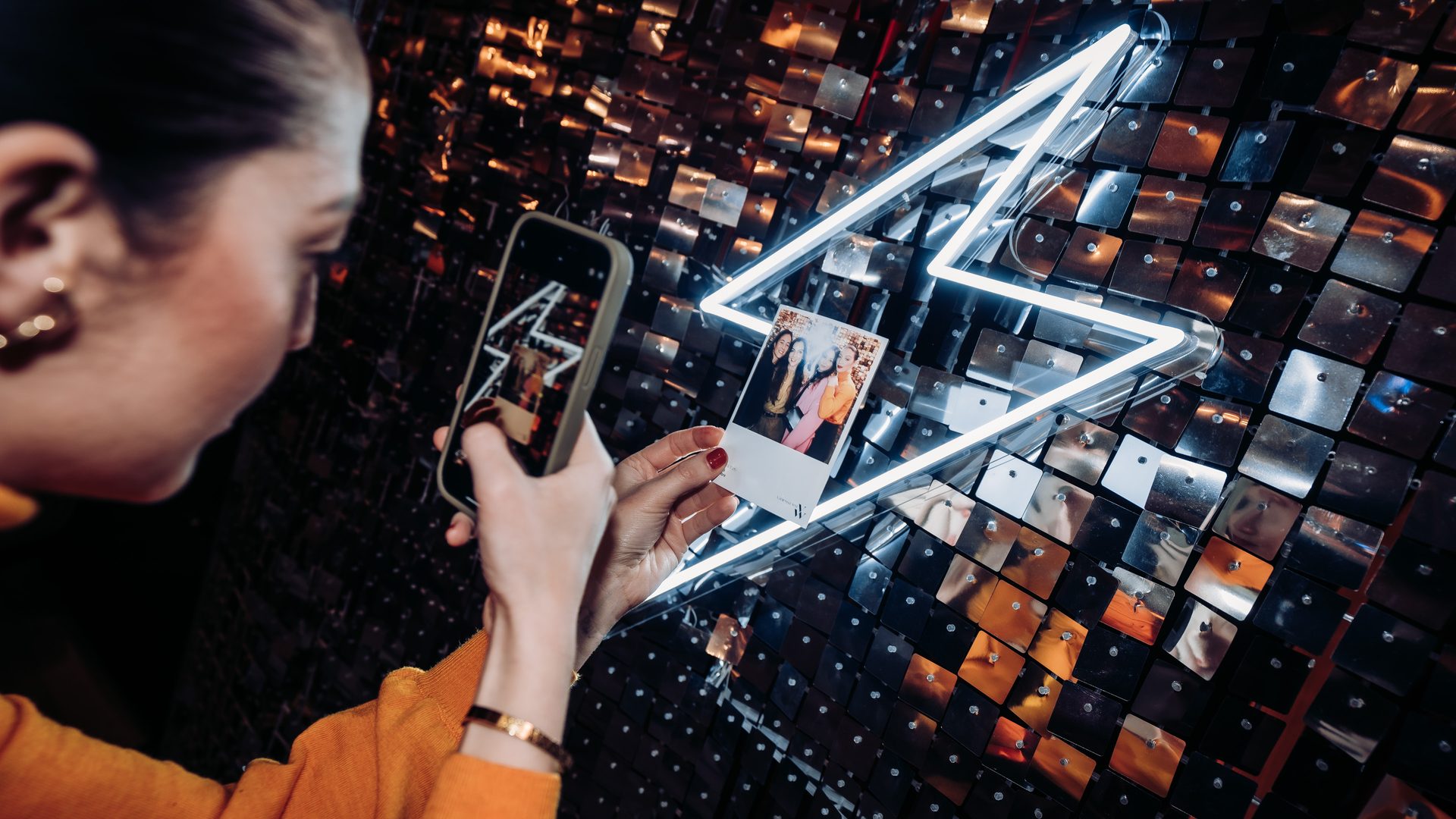 Invitée photographiant avec son téléphone son tirage photo souvenir devant un mur de sequins et un éclair en néon, lors de la soirée de lancement de Vanessa Wu organisée par Weevup