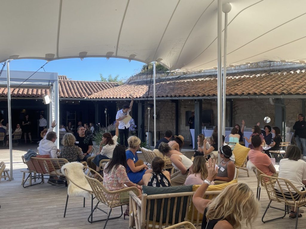 Un animateur debout, feuille en main, s’adresse aux participants d’Excelia, dont plusieurs lèvent la main, sous la tente du Bastion à Saint-Martin-de-Ré