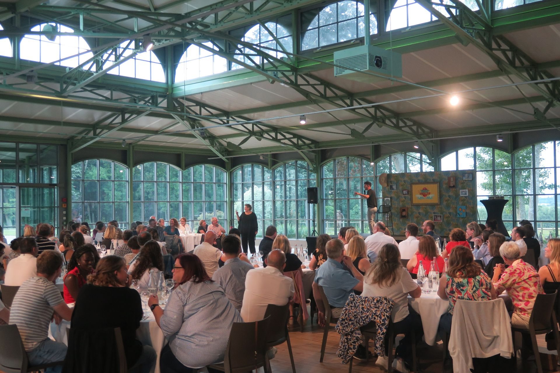 Invités de la soirée annuelle IGAM, organisée par Weevup, attablés autour de tables rondes nappées de blanc dans une grande verrière aux baies ouvertes sur les arbres