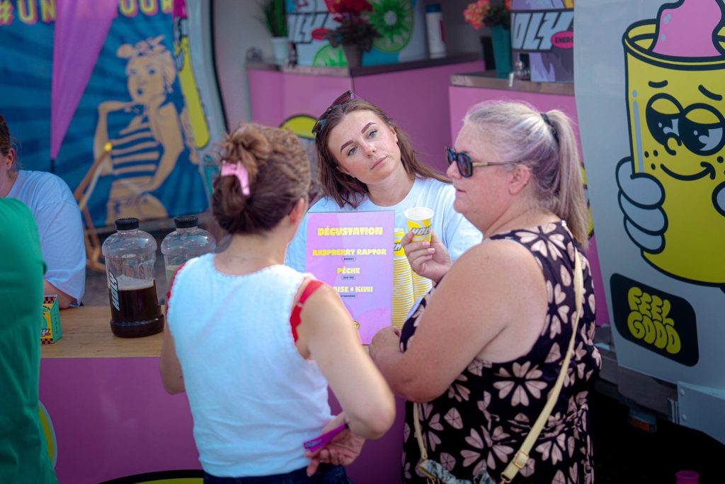 Hôtesse présentant la carte de dégustation Holy à deux festivalières au comptoir, au Lovely Brive Festival