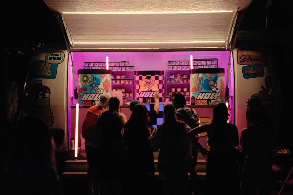 Comptoir de la caravane Holy éclairé en rose la nuit, présentoirs de produits et silhouettes de festivaliers