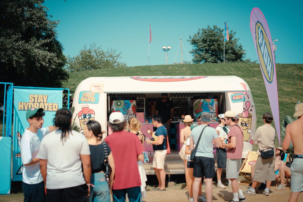 Caravane Holy en journée au Marvellous Island Festival, panneau Stay Hydrated et file de festivaliers au comptoir