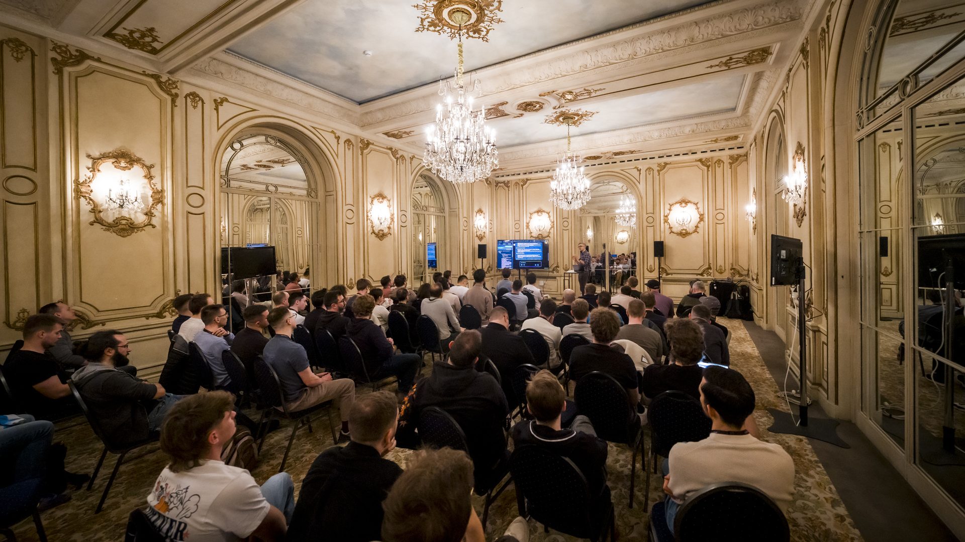 Plénière de la convention de janvier du Groupe Positive à Bruxelles, organisée par Weevup : participants assis face aux écrans dans un salon aux moulures dorées, sous des lustres en cristal
