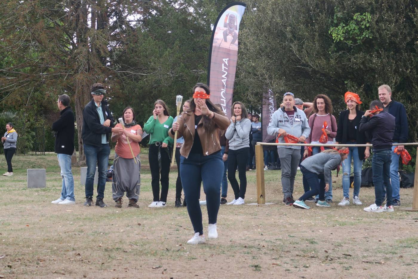 Participante aux yeux bandés d'un foulard orange avançant sur la pelouse d'un parc, sous le regard de son équipe, pendant la journée team building de Cerfrance organisée par Weevup
