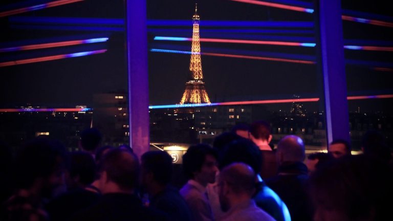 Invités de la convention 3DS Outscale devant une baie vitrée avec vue sur la tour Eiffel illuminée, à Paris, sous des néons bleus et rouges