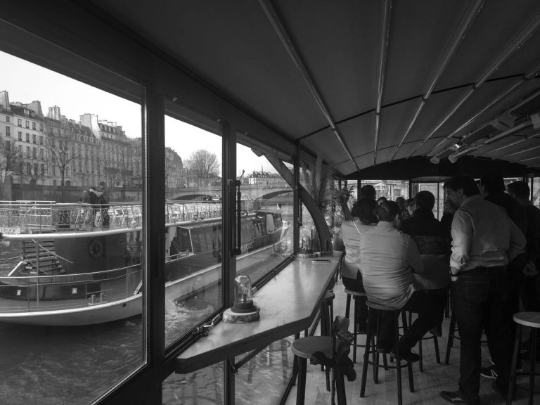 Invités de la SNCF debout au comptoir d’une péniche à quai sur la Seine, face aux immeubles parisiens (photo en noir et blanc)