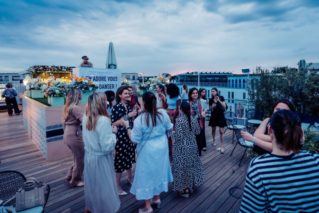 Invités de la cocktail party d’été de Dior réunis sur un rooftop à la tombée du jour, devant une cabine DJ fleurie