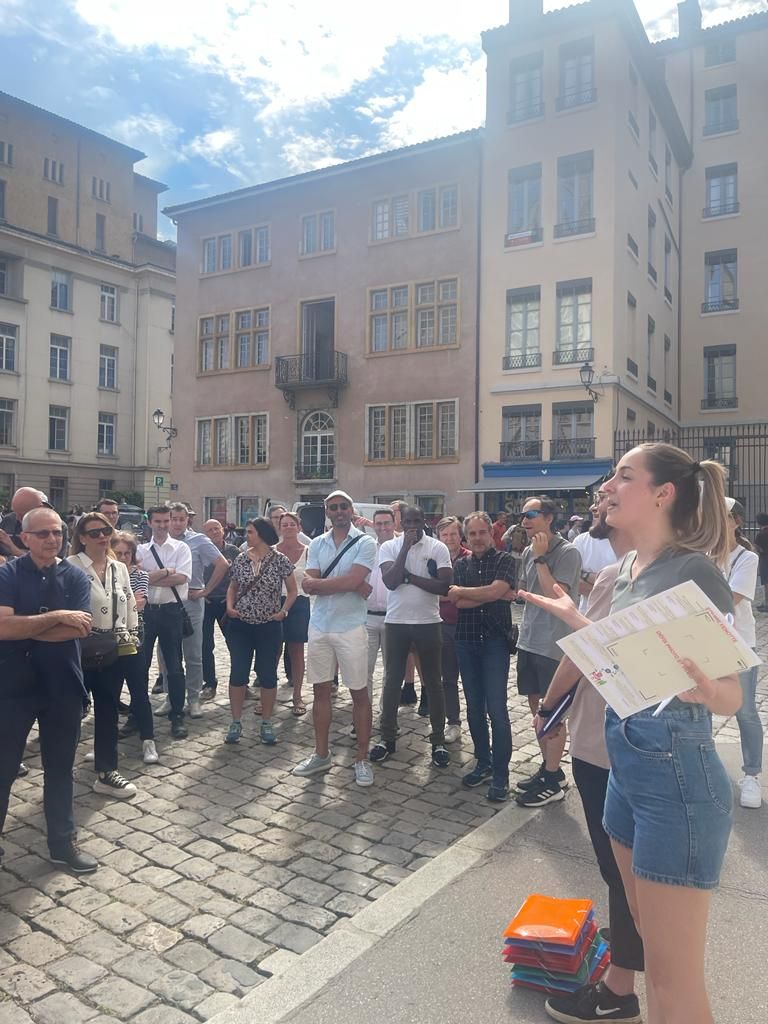 Briefing du rallye urbain sur une place pavée du Vieux Lyon : une animatrice donne les consignes, document en main, aux participants de Partecis
