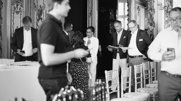 Invités de la journée clients Rimini Street debout près des rangées de chaises du salon de la plénière, à la Maison Ducasse Baccarat, documents et téléphones en main (photo en noir et blanc)