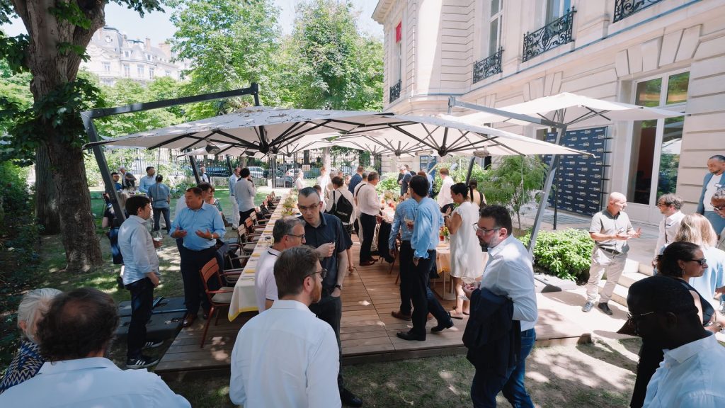 Apéritif dans le jardin de la Maison Ducasse Baccarat, invités verre à la main autour des tables dressées sous de grands parasols, photocall Street Smart au fond