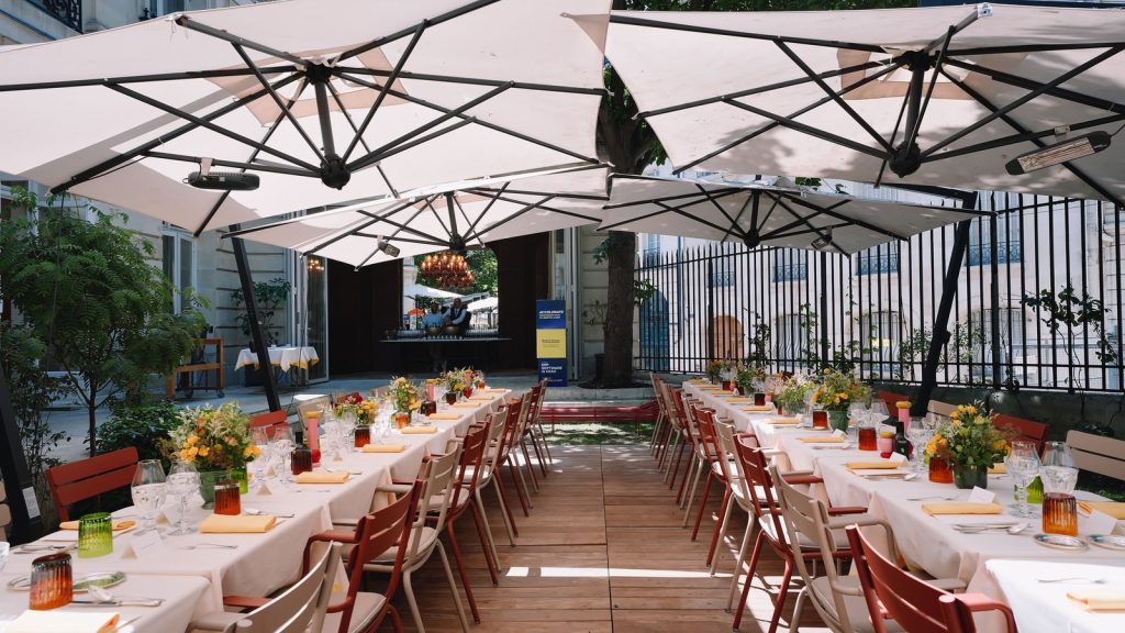 Tables du déjeuner dressées sous de grands parasols blancs sur la terrasse en bois du jardin de la Maison Ducasse Baccarat, avec bouquets de fleurs et chaises rouges et beiges