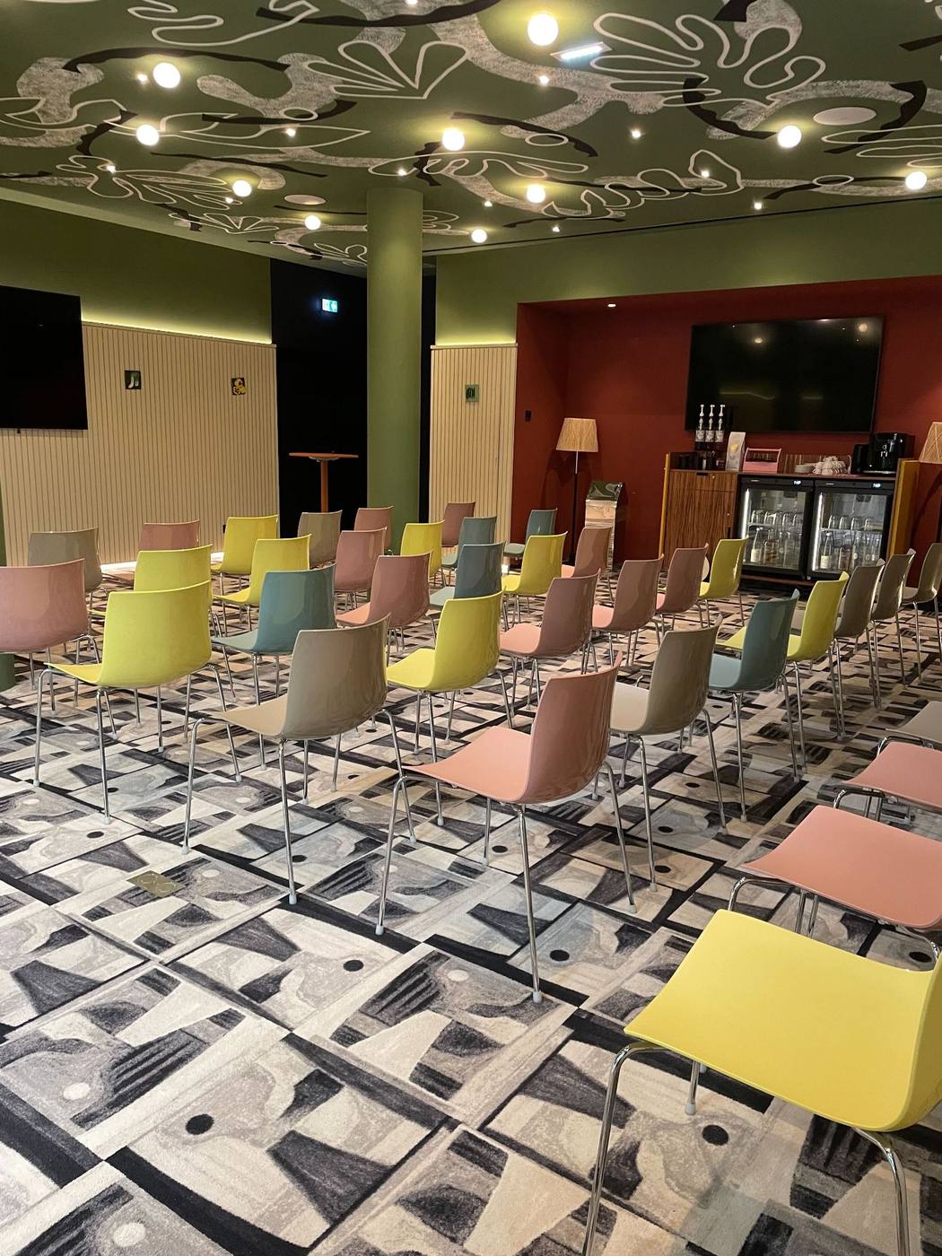 Salle aux chaises colorées installée en rangs face aux écrans, sous un plafond vert à motifs, prête à accueillir une étape du Parthena Roadshow 2025 organisé par Weevup
