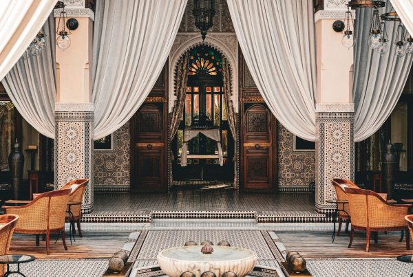 Patio d'un palais de Marrakech aux voilages blancs, colonnes en zellige, portes en bois sculpté et fauteuils en rotin autour d'une vasque en marbre, cadre d'un séminaire d'entreprise au Maroc