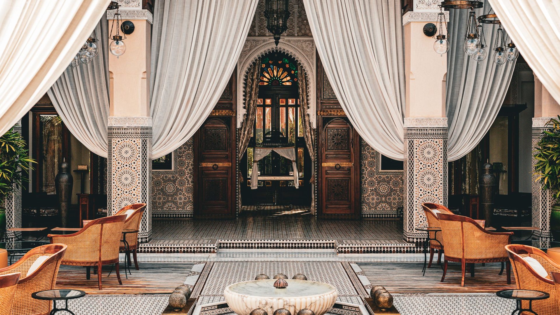 Patio d'un palais de Marrakech aux voilages blancs, colonnes en zellige, portes en bois sculpté et fauteuils en rotin autour d'une vasque en marbre, cadre d'un séminaire d'entreprise au Maroc