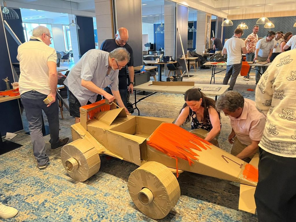 Collaborateurs de Partecis assemblant au ruban adhésif une Formule 1 en carton décorée d’orange, dans un salon du Radisson Blu Paris Marne-la-Vallée