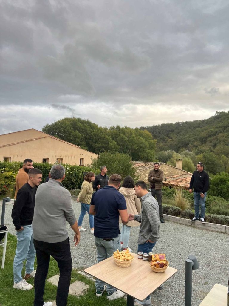 Collaborateurs de Pacific réunis pour un apéritif en plein air autour d’une petite table, devant une bâtisse en pierre et des collines boisées, au domaine de Ribiera en Provence