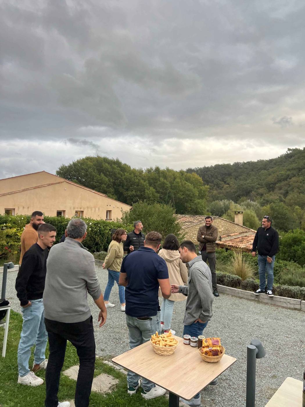 Collaborateurs de Pacific réunis pour un apéritif en plein air autour d'une petite table, devant une bâtisse en pierre et des collines boisées, au domaine de Ribiera en Provence