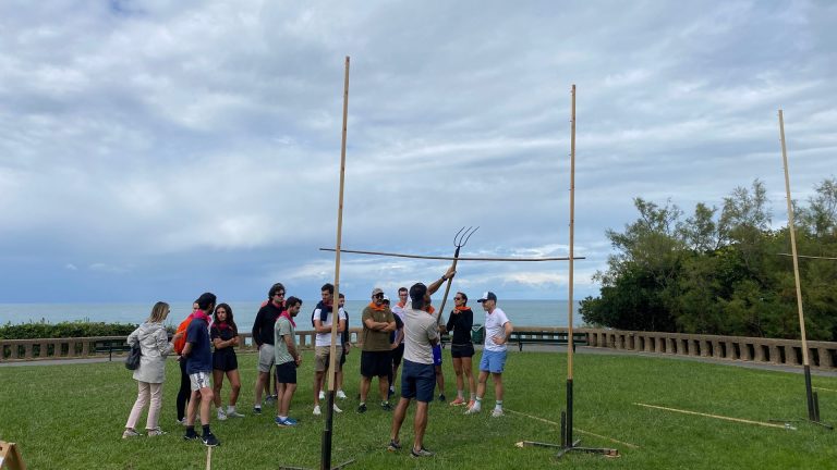 Libon incentive trip participants, orange scarves round their necks, watching a facilitator demonstrate a Basque games event with a pitchfork, on a lawn facing the ocean in Biarritz